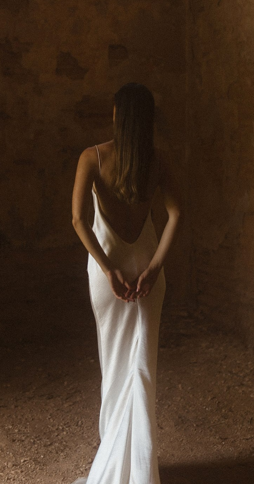 Woman in a white backless dress stands in a dimly lit room, facing a textured wall with hands clasped behind her back.
