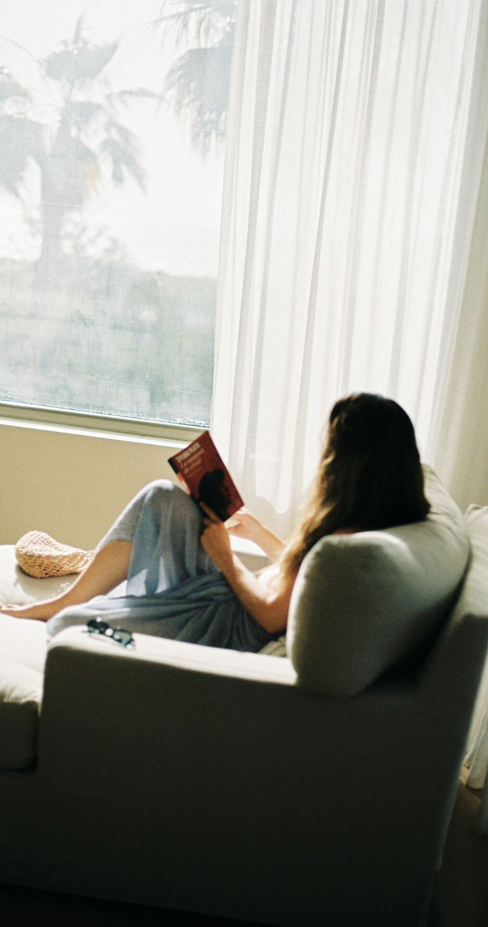 Woman sitting on a couch, reading a book by a bright window with sheer curtains. A hat and sunglasses are beside her.